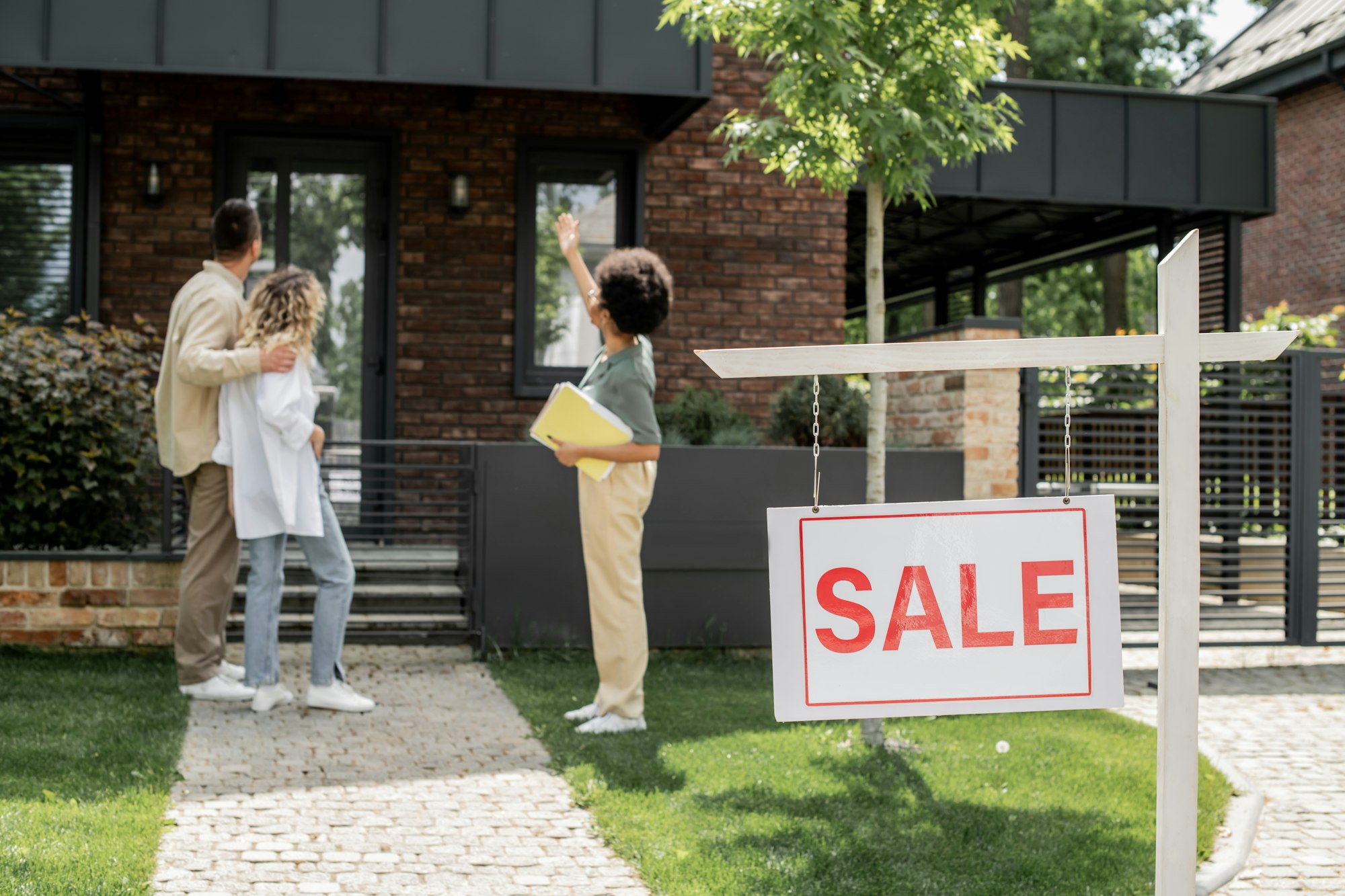 african american real estate agent showing house to hugging couple near for sale signboard